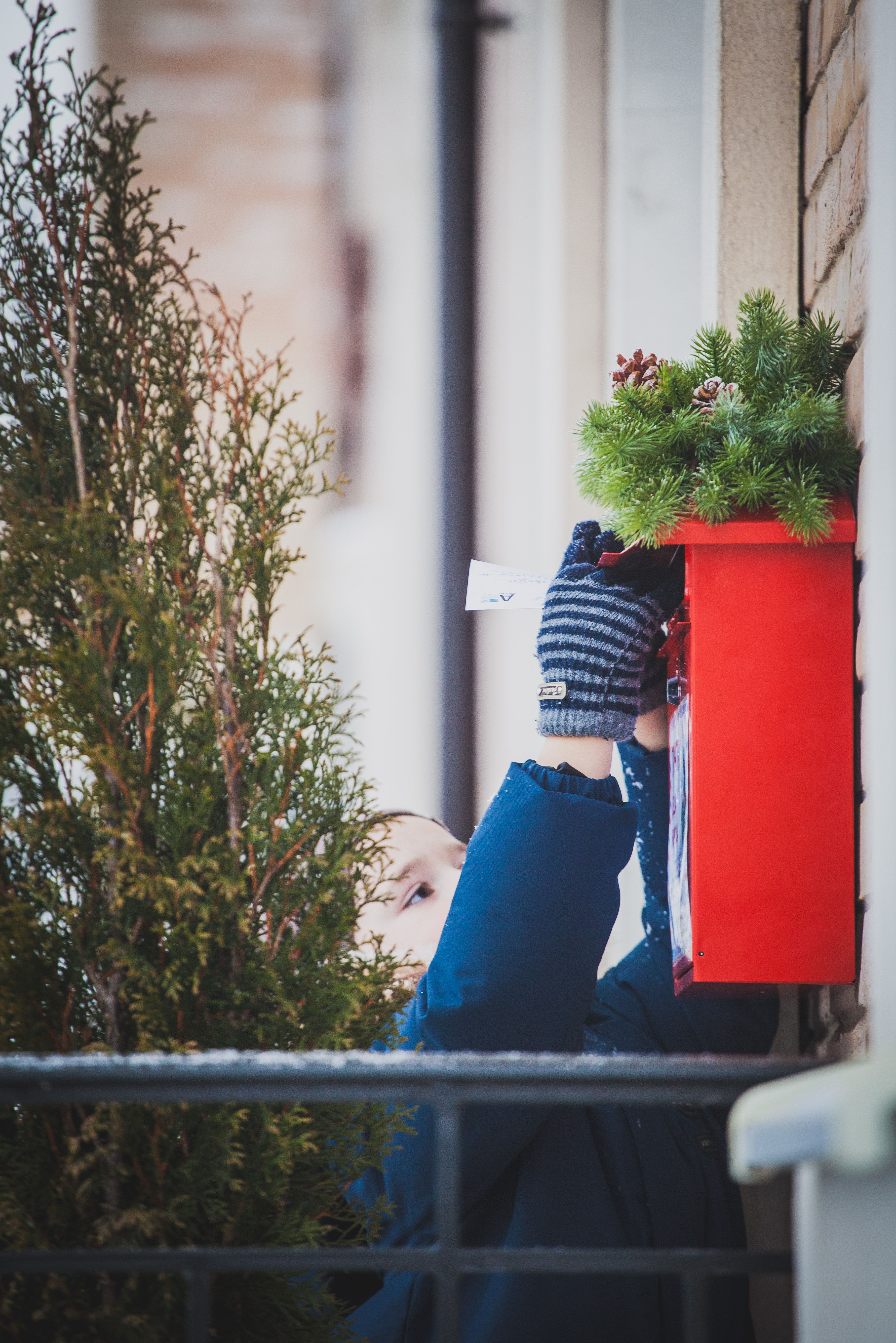 A child reaches up to place a letter in a red Christmas mailbox adorned with festive decorations.