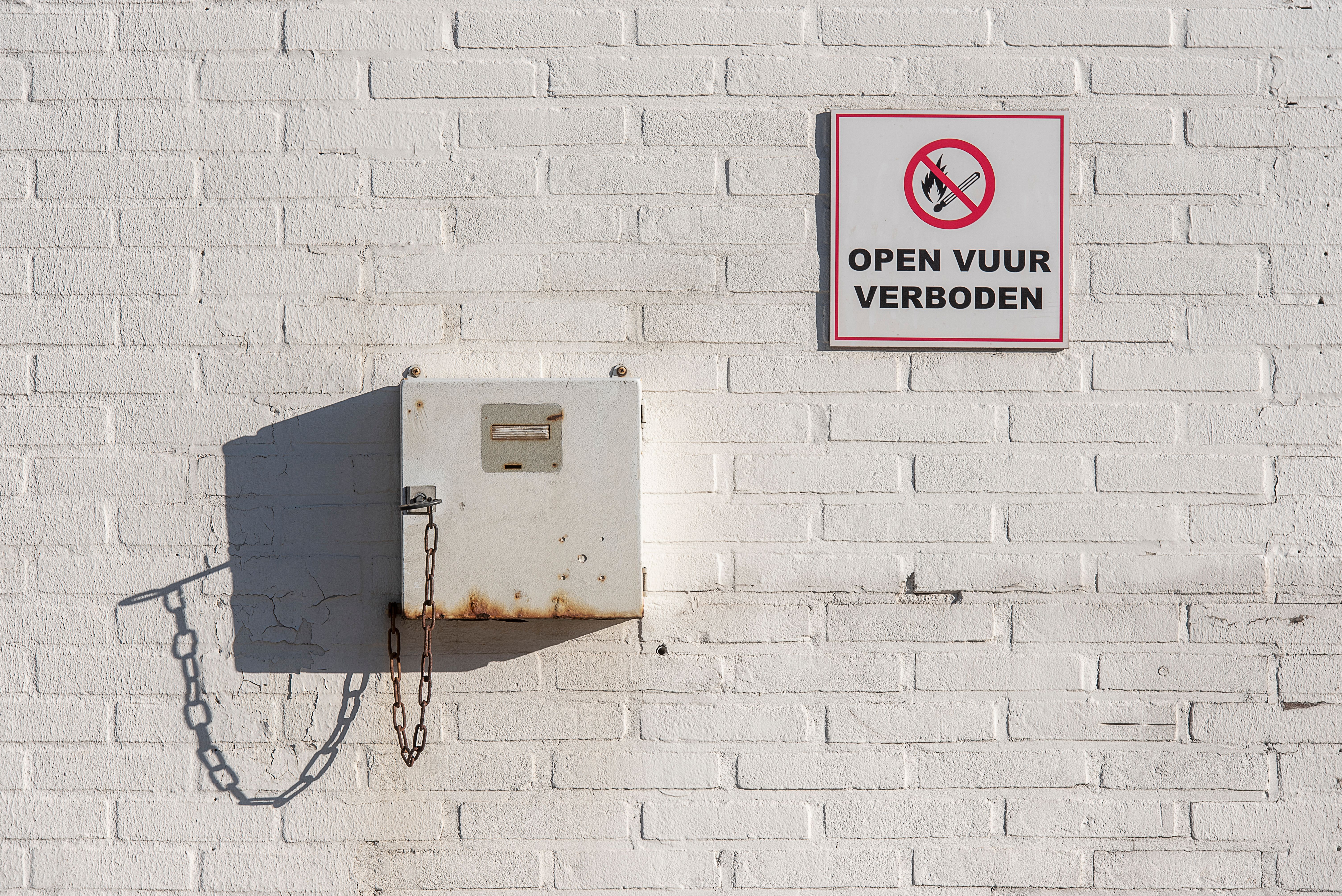 White wall with a no open fire sign and metal box with chain, casting shadow.