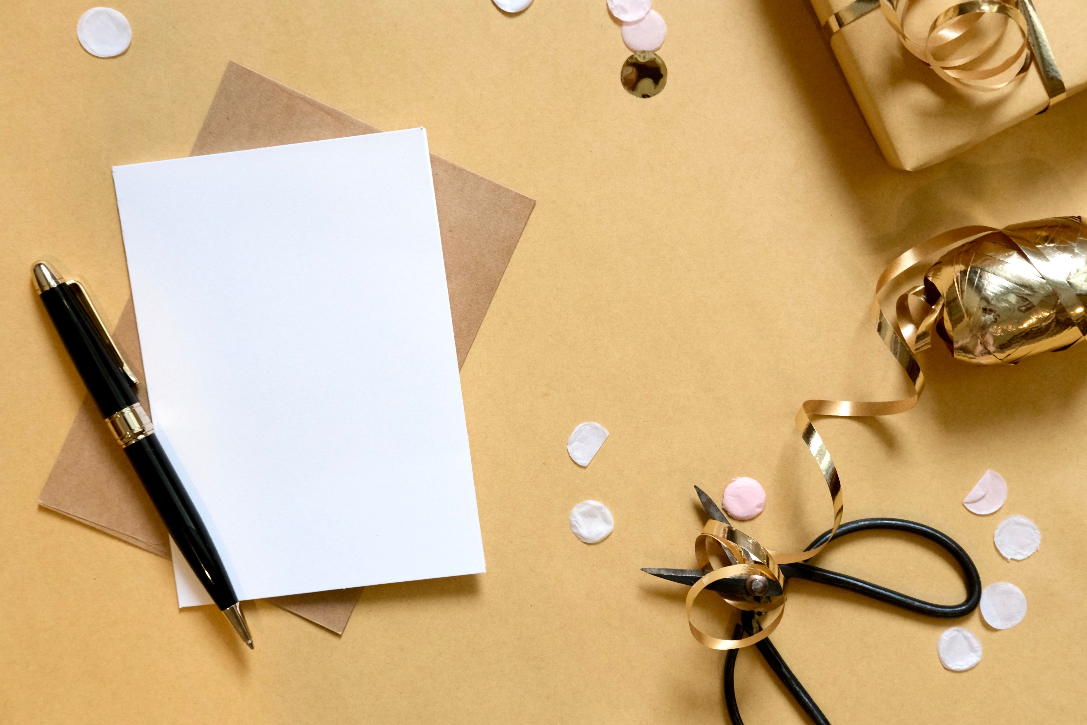 Flat lay of a blank card, pen, scissors, and gift-wrapping items on a brown surface.