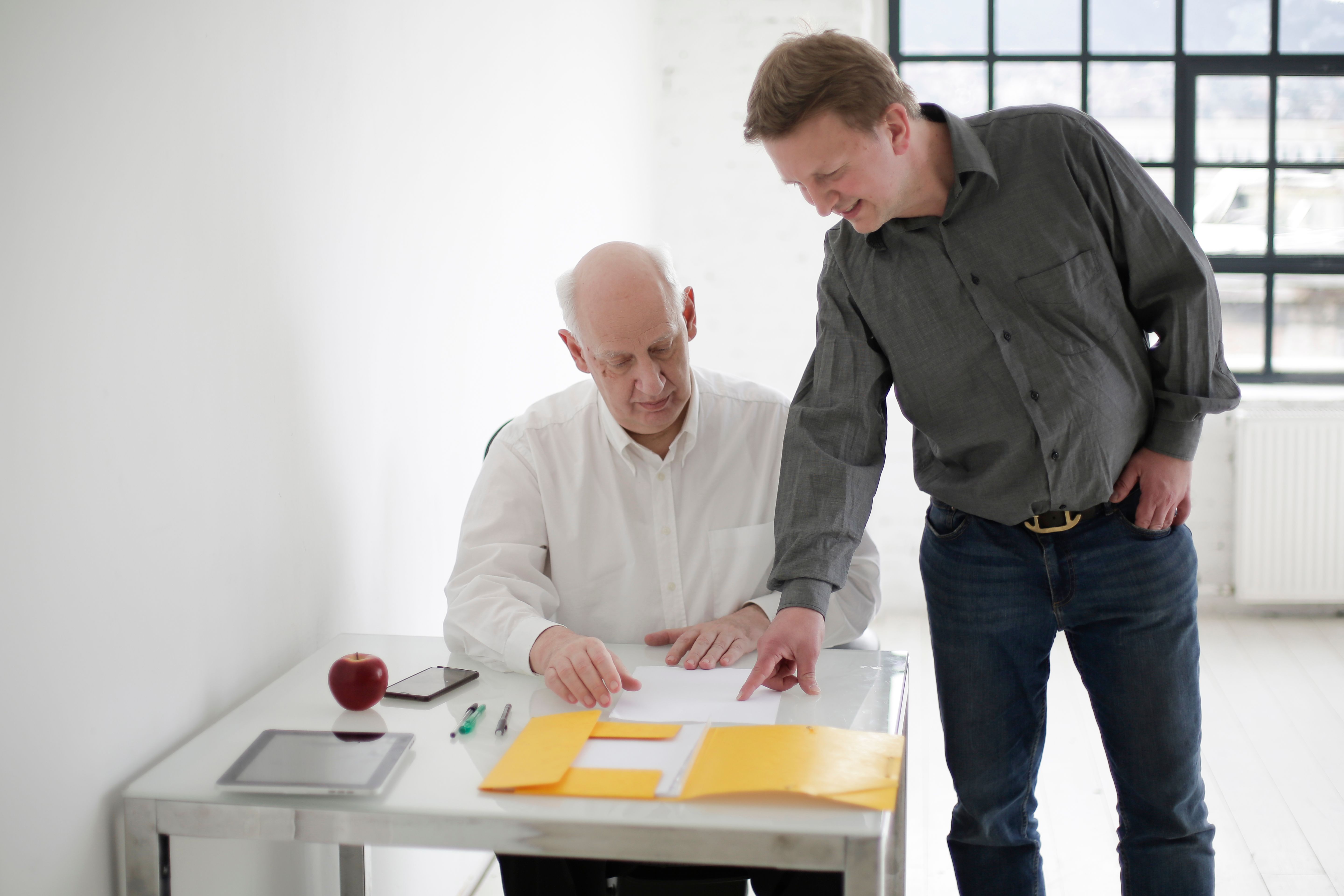 Positive young guy in casual wear showing documents to aged bald boss in white cozy workspace