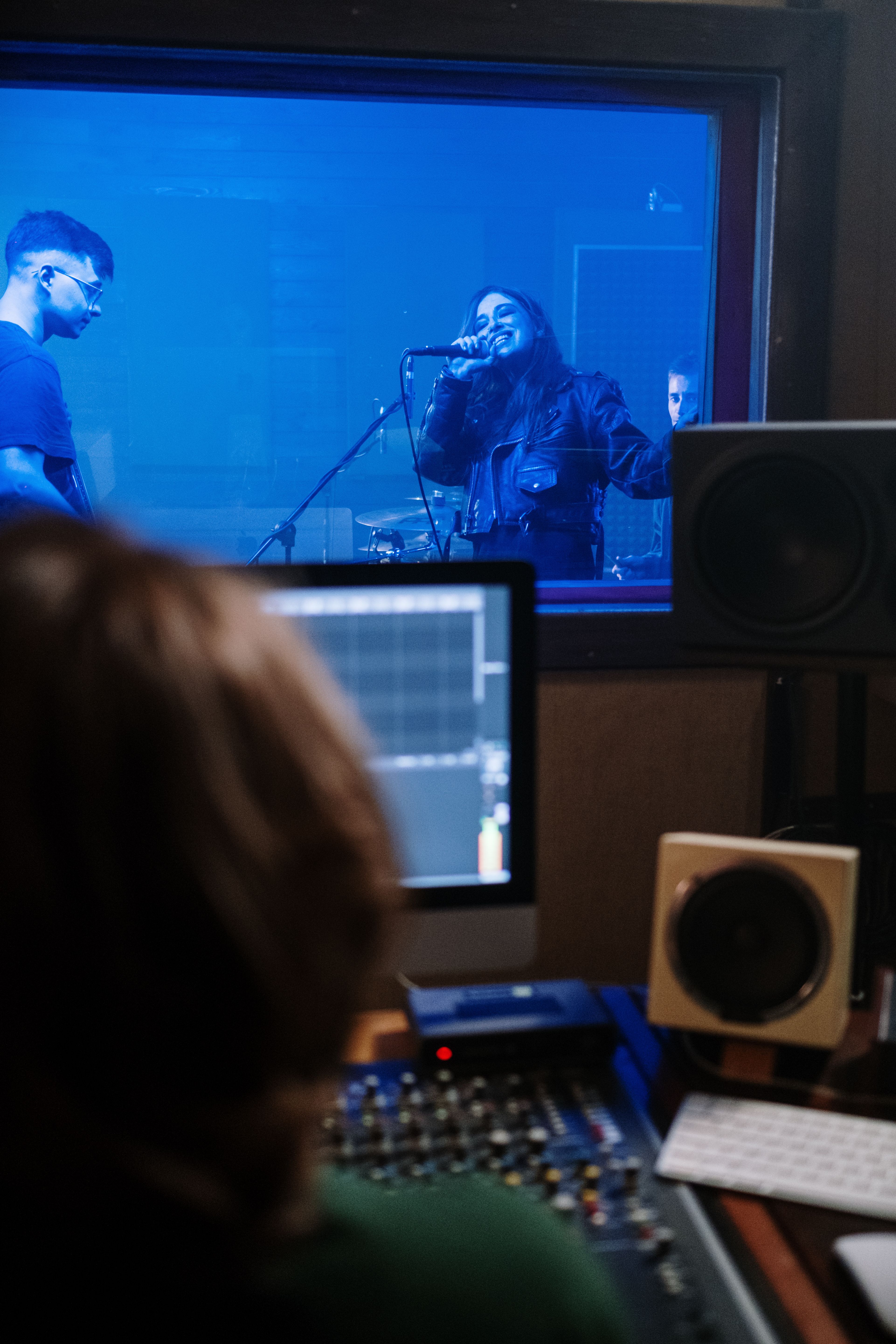 Musicians performing in studio with blue lighting, captured from control room perspective.