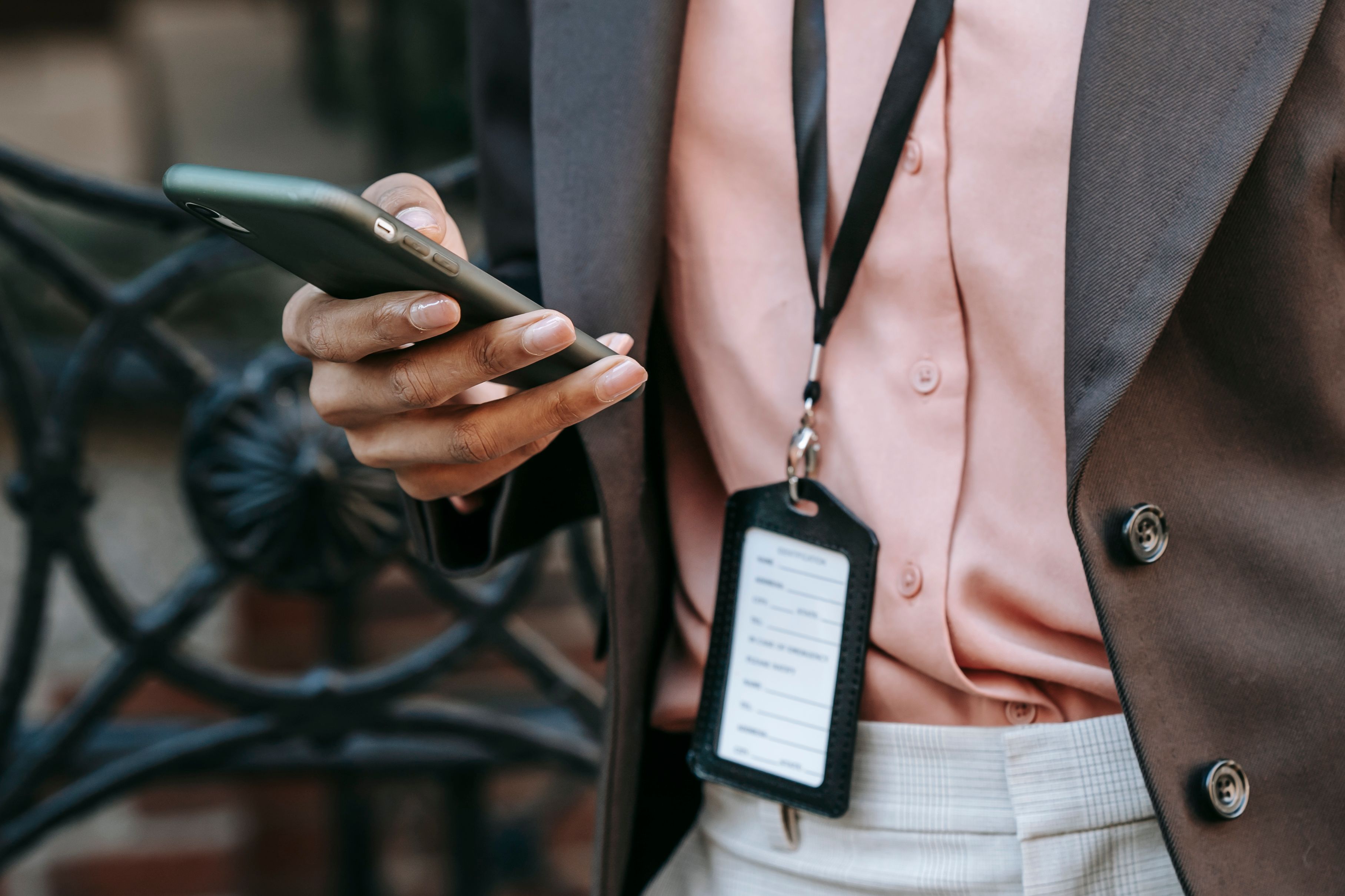Crop unrecognizable African American female entrepreneur in stylish suit with name tag messaging on mobile phone while standing on city street in daytime