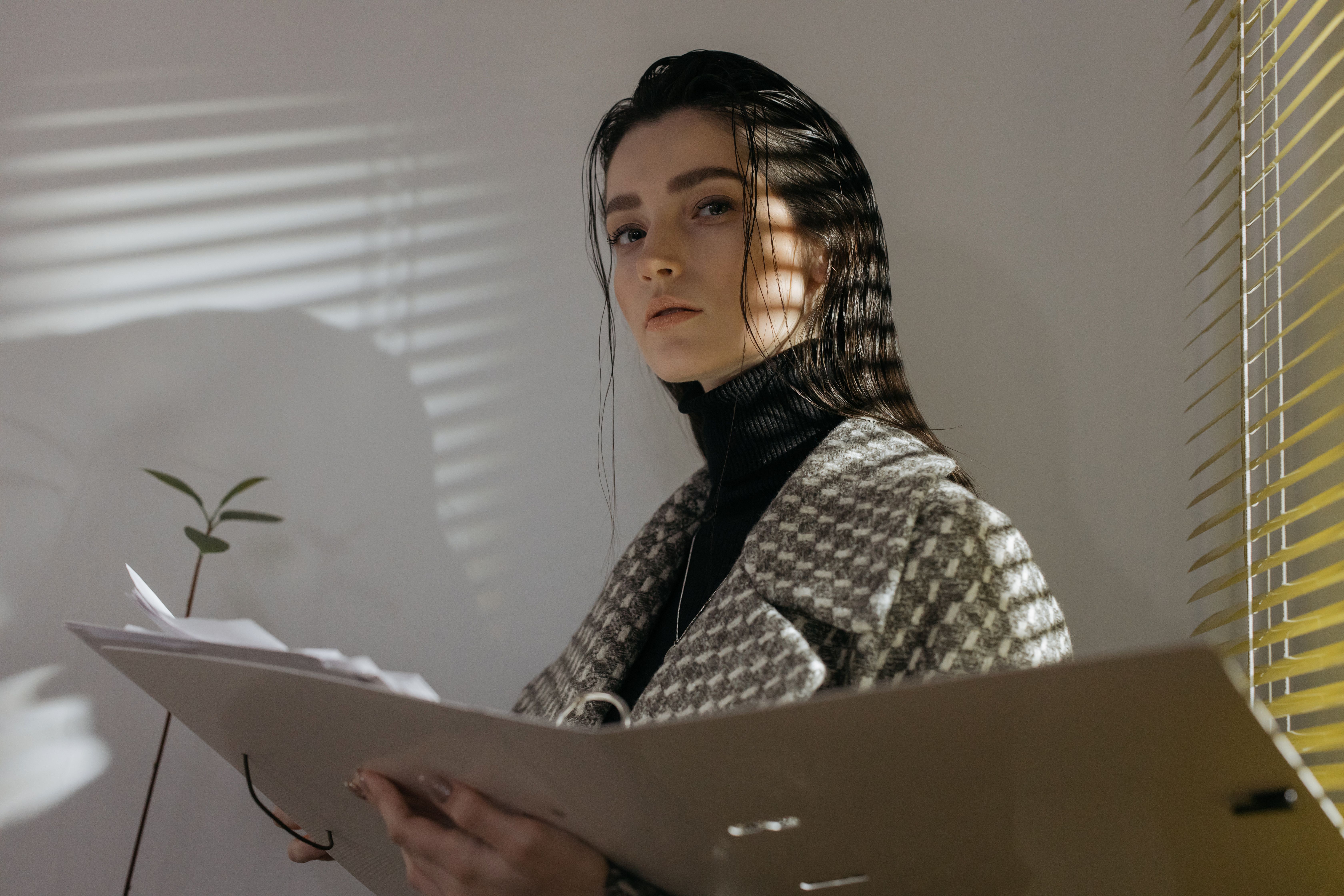 Stylish woman in houndstooth coat holding file folder. Light and shadow create dramatic effect.