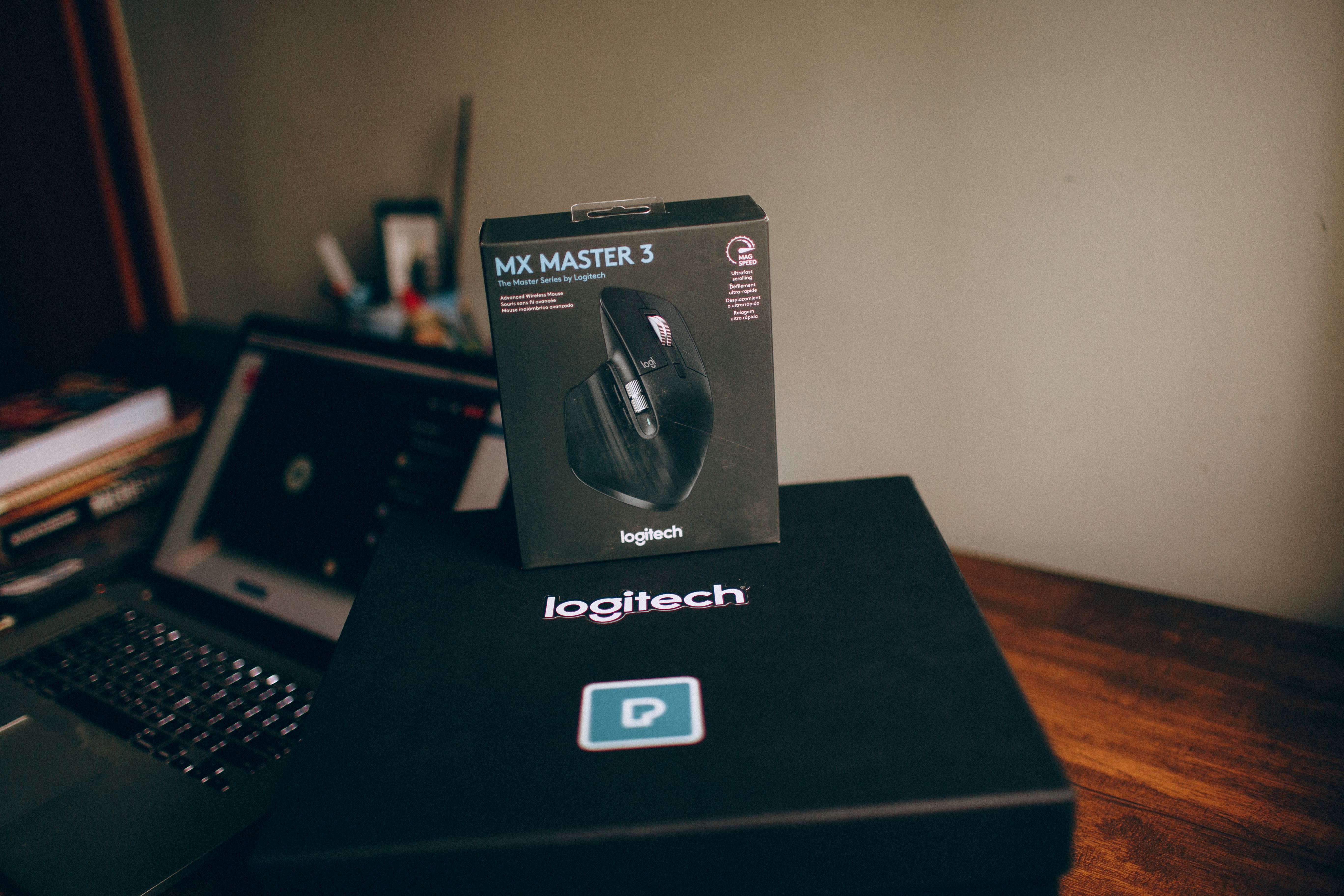 Image of Logitech MX Master 3 wireless mouse box on a desk with a blurred laptop in background.