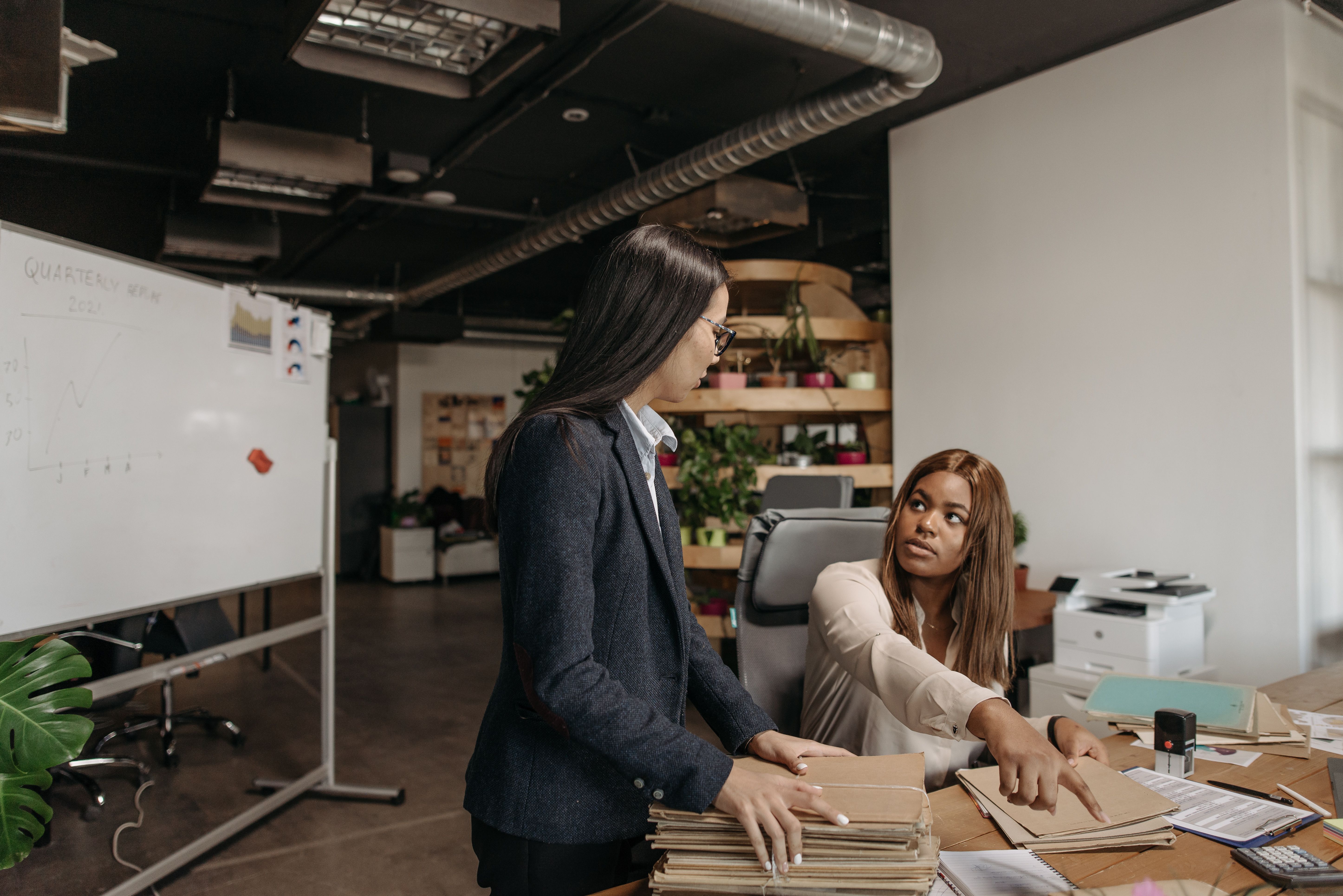 Two professional women discussing work in a modern office setting with documents and a whiteboard.