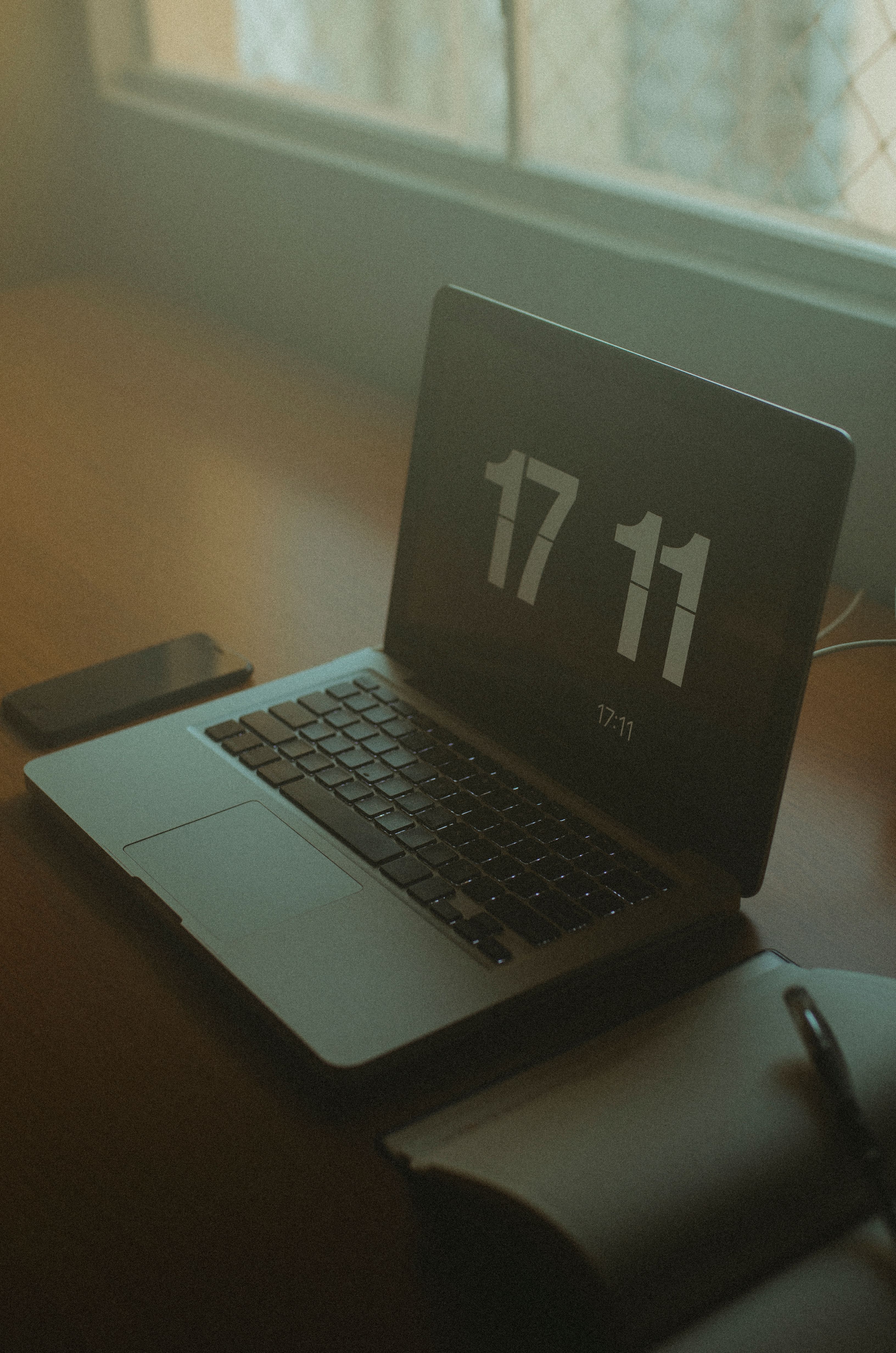 Minimalist workspace featuring a laptop displaying digital time, an open notebook, and natural light from window.