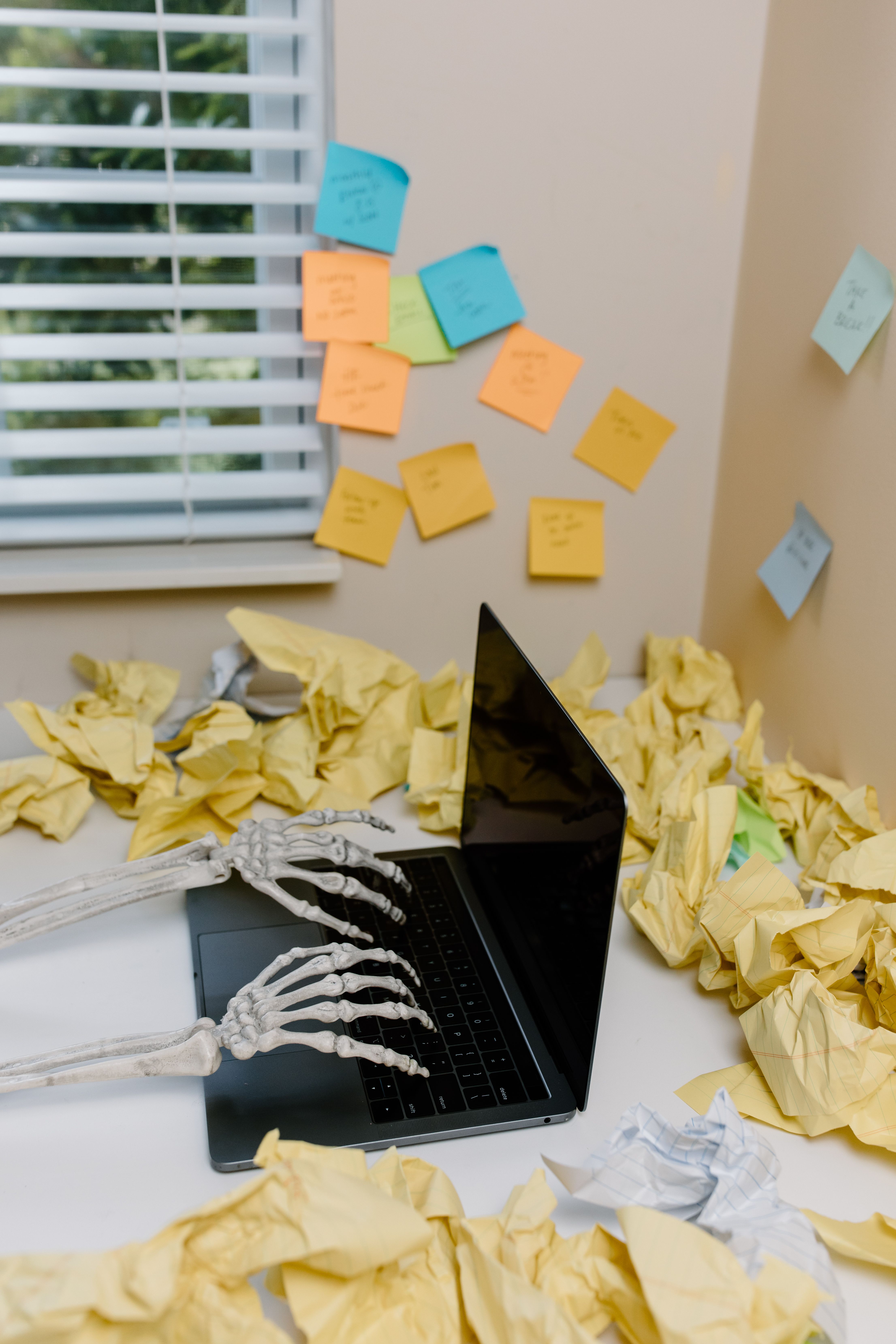 Creative depiction of a skeleton working on a laptop amidst cluttered papers in an office setting.