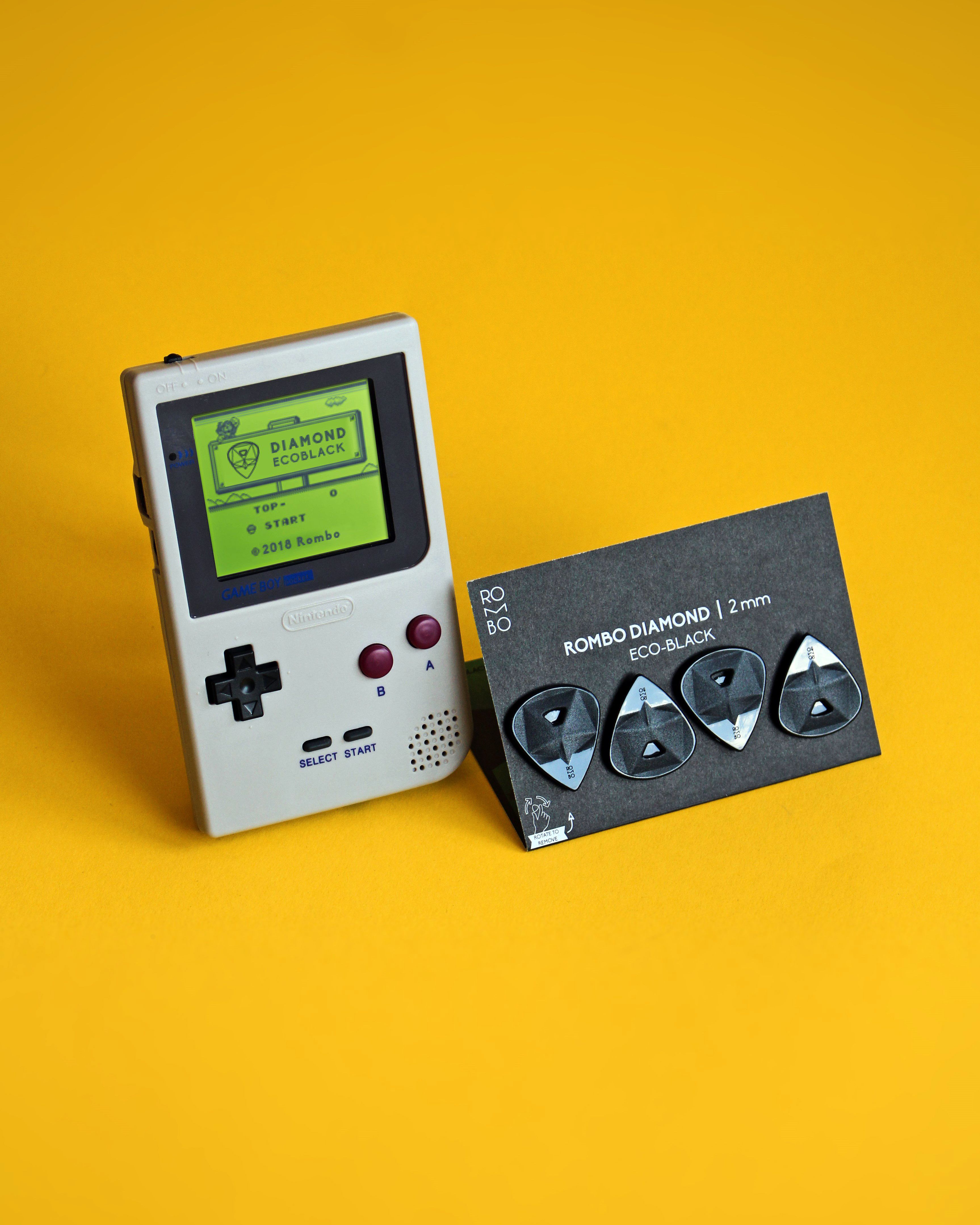 A classic Game Boy and Rombo Diamond eco-black guitar picks on a bright yellow backdrop, showcasing retro gaming and music accessories.